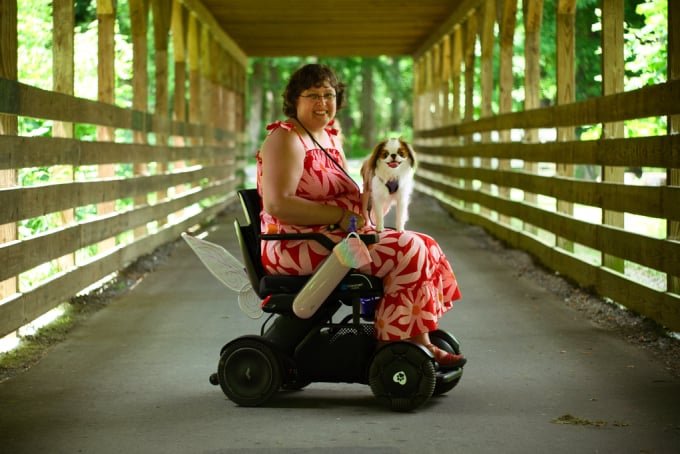 A white woman with short brown hair sits in a power wheelchair with wings on the back.  The wheelchair is positioned sideways in the middle of a covered bridge.  A sable and white Japanese Chin sits on her lap.  They are both looking at the camera and smiling.