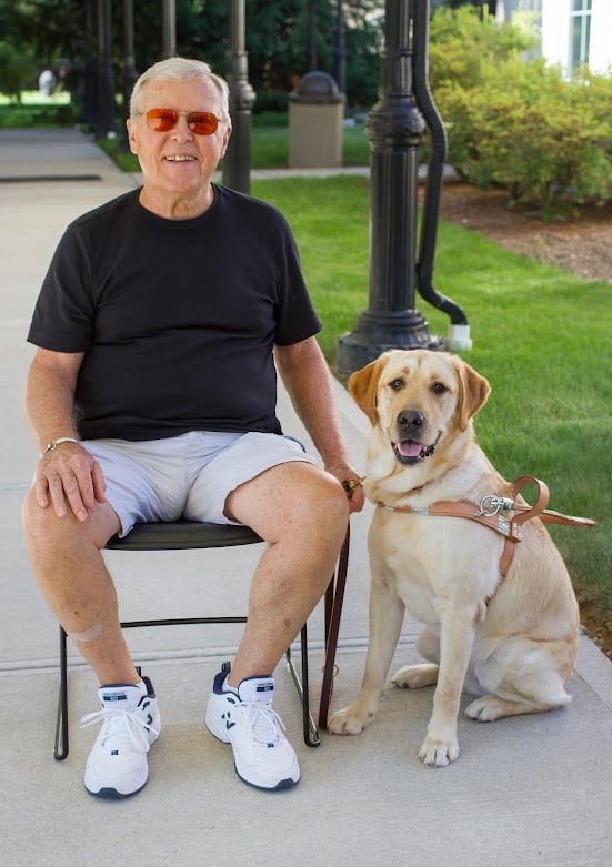 A smiling white man with short gray hair and sunglasses sits outdoors in a folding chair wearing a black t-shirt and white shorts with tennis shoes. His guide, a yellow Labrador Retriever in harness, sits at his side.