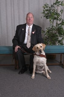 A smiling white man with short brown hair and wearing a suit and tie sits on a blue bench in a lobby with his guide, a cream-colored labrador retriever in harness, seated on the floor next to him.